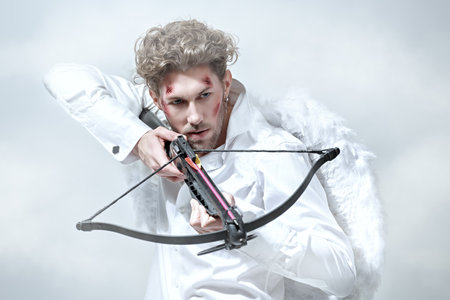 A warlike handsome man, the God of love Cupid with wounds on his face, intends to shoot an arrow at his victim. Fantasy. Valentine's Day. White background with clouds.の写真素材