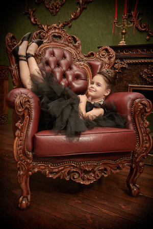 Full-length portrait of a pretty little girl dressed in a puffy black dress and with evening hairstyle and jewelry lies on a vintage armchair and smiles. Luxury apartments. Evening Fashion for girls.の写真素材