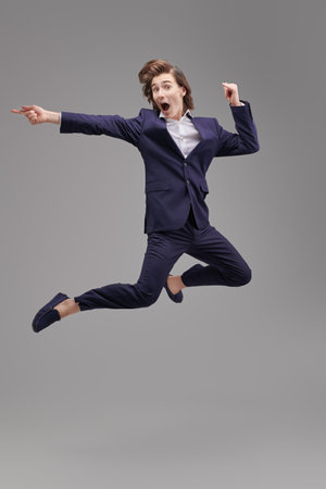 An excited teenage boy in a classic suit with white shirt makes a big jump with all his might. Full length portrait on a gray studio background. Modern education. Striving for success, big ambitions.の写真素材