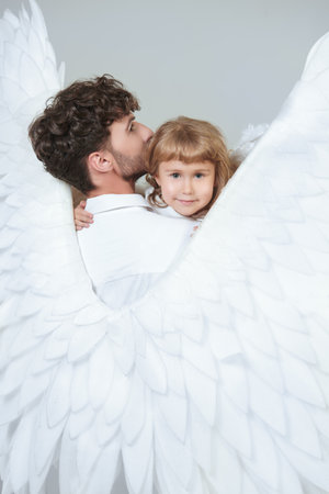 A male guardian angel holds a little baby girl in his arms, covering her with his wings. White studio background.の写真素材