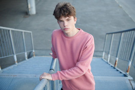 Youth fashion. A modern young man fashion model poses in a pink sweatshirt on a metal staircase near a shopping mall. Urban style.の写真素材