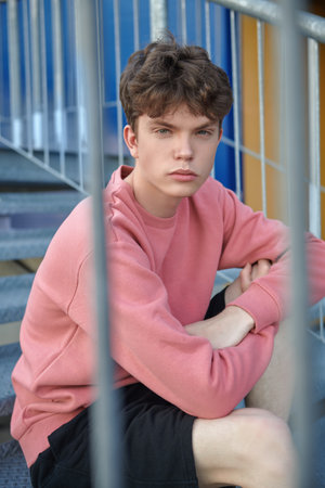 A modern young man fashion model poses in a pink sweatshirt and black shorts on a metal staircase. Urban style. Youth fashion.の写真素材