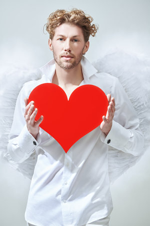 Sexy handsome male Cupid angel in white clothes and with white wings holding a heart. Skylight in the background. God of love Cupid. Valentine's Day.の写真素材