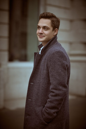 A handsome modern young man in a coat stands on a city street on a cloudy day and smiles. People and emotions, lifestyle.の写真素材