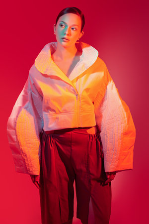 Fashion and Style. Beautiful fashion model girl posing in a designer white jacket with wide sleeves and brown trousers with wide legs. Studio shot against a red background in mixed color light.の写真素材