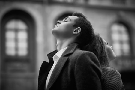 Black and white portrait of a man and a girl standing with their backs to each other, eyes closed and lost in their thoughts against the backdrop of a cloudy city. People, feelings and relationships.の写真素材