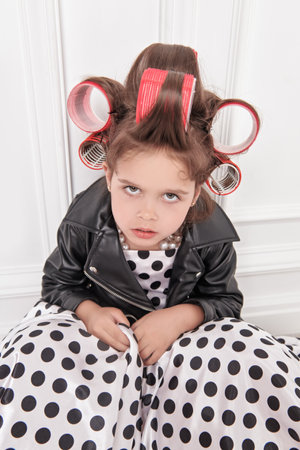 A little fashionista girl in a smart dress with polka dots, in a leather jacket and with curlers on her head is going to a party and unhappy with something. Children's Fashion. Pin-up style.の写真素材