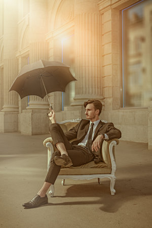 A well-groomed young man in a black suit with a white shirt and tie sits in an expensive chair on a city street under a black umbrella covering from sun. Business concept. Security and confidence.の写真素材