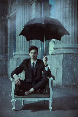 A well-groomed young man in a black suit with a white shirt and tie sits in an expensive chair on a city street with rain under a black umbrella over his head. Business concept. Security and confidence.の写真素材