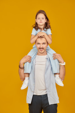 Family portrait of a bored little daughter sitting on her happy dad's neck. Studio shot on a yellow background.の写真素材