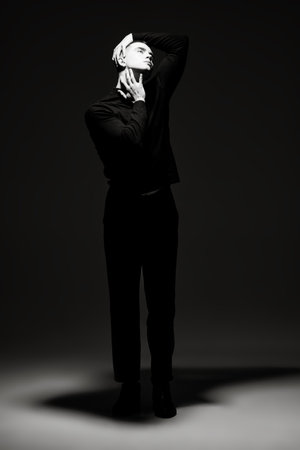 Art portrait of a young man in black clothes, who stands in the dark and a beam of light illuminates his face. Full-length black and white portrait against a black background. Psychology. Theater.の写真素材
