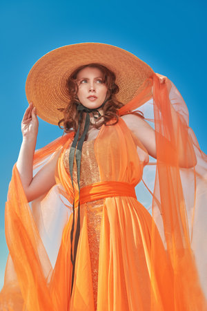 Summer fashion. Portrait of a gorgeous lady in a light orange evening dress and a wide-brimmed straw hat against a blue sky. Elegant summer style.の写真素材