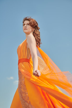 Summer fashion. Beautiful girl fashion model posing in a flying orange dress against a blue sky. Bright colors of summer.の写真素材