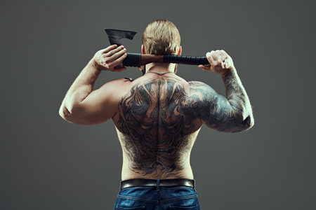 Courage and physical strength. A handsome muscular tattooed man stands with his back with a large ax in his hands, posing confidently. Grey studio background.の写真素材