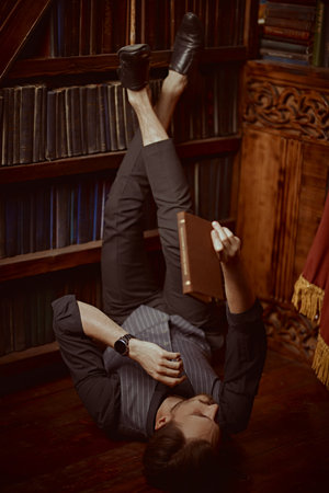 A handsome, well-dressed man with dark hair and a beard relaxes on the floor in a home library with...