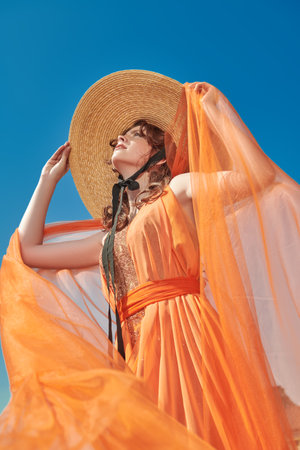 Summer women's fashion. Portrait of a gorgeous lady in a light orange evening dress and a wide-brimmed straw hat against a blue sky. Elegant summer style.の写真素材