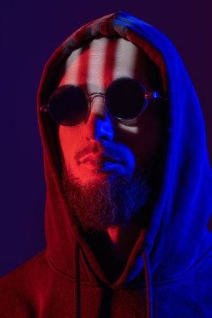 A brutal shaven-headed bearded man with a lip piercing and wearing black sunglasses and black hood....