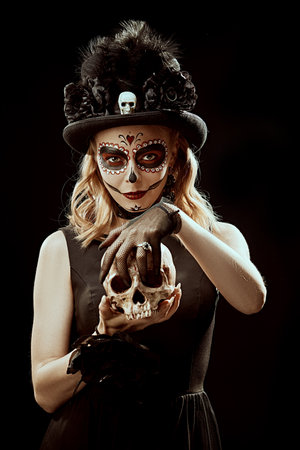 Art portrait of a Sugar Skull girl posing in a carnival headwear and with a human skull in her hands. Day of the Dead. Dia de los Muertos. Halloween. Calavera Catrina.の写真素材