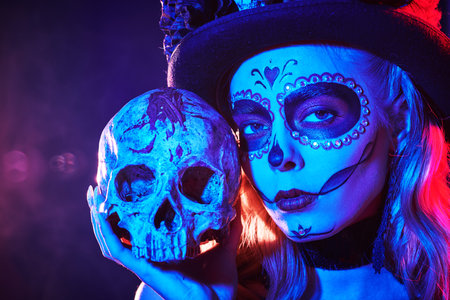 Calavera Catrina. Art portrait of a Sugar Skull girl posing in a carnival headwear and with a human skull in her hands in festive colorful lighting. Day of the Dead. Dia de los Muertos. Halloween.の写真素材