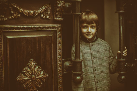 A smiling little boy in a retro style coat and cap stands at doors, smiles and looks at the camera. Vintage background. Children's fashion, retro style. Copy space.の写真素材