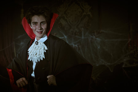 An attractive young vampire in an elegant tailcoat and a cloak stands in an old castle covered in cobwebs and smiles slyly. Vampires novel. Copy space. Halloween.の写真素材