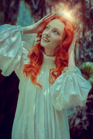 The heroine of the novel. A beautiful red-haired girl princess in a long white shirt with a crown on her head stands in the forest. Romanticism of the 18th and 19th centuries. Fantasy.の写真素材