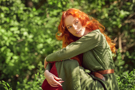 Happy romantic girl with magnificent red hair in a historical celtic dress sits in a sunny forest and smiles. Historical reconstruction of Middle Ages. Fantasy heroine.の写真素材