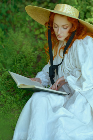 A beautiful romantic girl with charming wavy red hair, dressed in a long white shirt and a wide-brimmed straw hat, rests with a book in a park on a sunny summer day. The era of romanticism, 18-19 centuries.の写真素材
