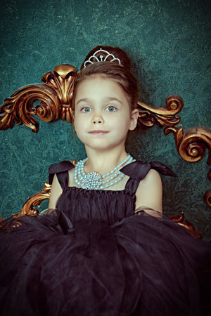 Evening hairstyles. A beautiful little girl model in a puffy black evening dress and an expensive pearl necklace poses in a luxury interior. Evening children's Fashion - clothing and jewelry.の写真素材