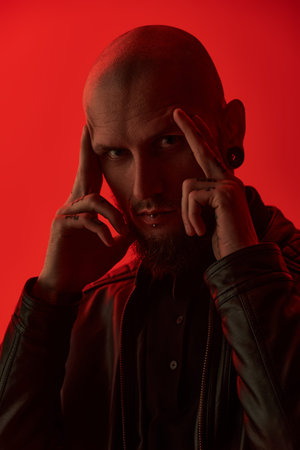 An informal skinhead man with a brutal appearance poses against a red studio background.