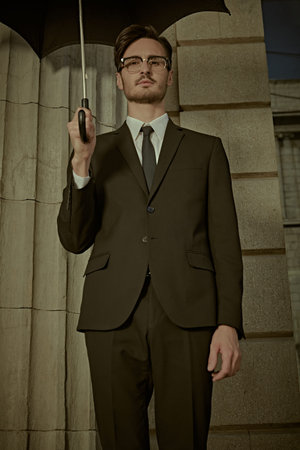 Businessman. Men's Fashion. A handsome young man in a classic black suit with a white shirt, a tie and with a black umbrella in his hand poses at the columns on a city street.の写真素材