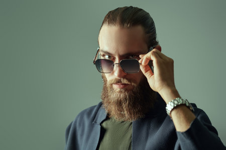 Style and Fashion for men. Portrait of a handsome good-looking man with a beard and mustache posing in a stylish knitted cardigan and fashionable sunglasses on a gray studio background. Copy-space.の写真素材