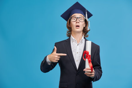 A modern graduate student in a suit and academic hat is surprised with his diploma. High quality education. Successful future. Blue studio background with copy space.の写真素材