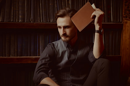 Rich lifestyle. A handsome, well-dressed man with dark hair and a beard sits in a home library with a book in a rich vintage interior.の写真素材