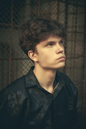 A modern young man in black clothes stands against a metal grid in a dark alley of a big city. Urban lifestyle.の写真素材
