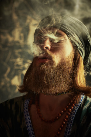 Lifestyle. A rastaman hippie looking man with a beard and long hair smokes a cigarette. Grunge background.の写真素材