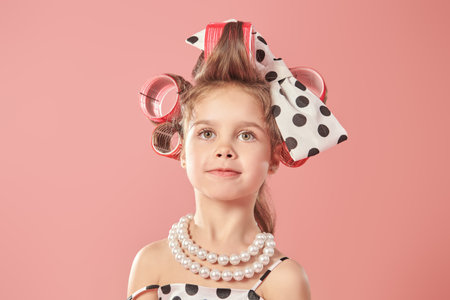 Fashion for kids - clothes and accessories. A little pretty girl in an elegant polka-dot dress and curlers on her head is going to a party. Pink studio background. Pin-up style.の写真素材