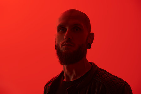 An informal skinhead man with a brutal appearance poses against a red studio background. Space for text.の写真素材