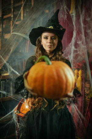 Halloween time. Pretty young witch with long curly hair in a black dress and hat holds joyfully a...