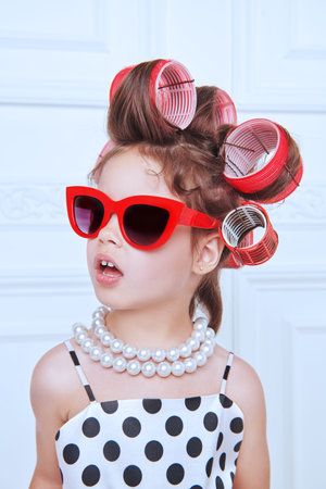 A little pretty girl in an elegant polka-dot dress, sunglasses and curlers on her head is going to a...