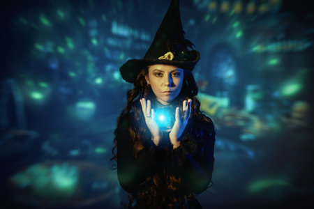 Magic. A charming sorceress in a black hat and dress conjures with a help of a shining crystal ball. Magical background. Halloween fairy tales.の写真素材