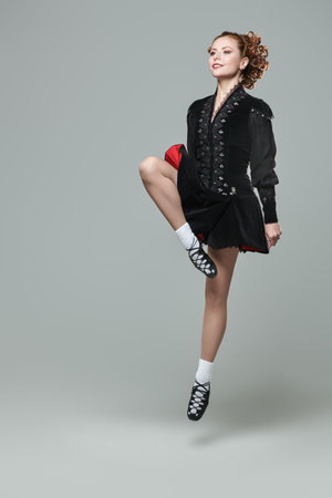 Traditional Irish dances. Professional dancer woman in national Irish costume makes a jump. Studio full-length portrait on a gray background. Culture.の写真素材