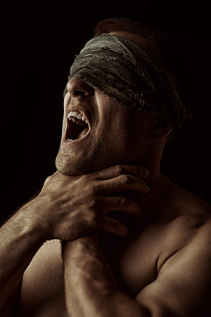 Psychological picture. A large, strong man with a naked torso stands with a gauze bandage over his eyes and screams. Studio portrait on a black background.の写真素材