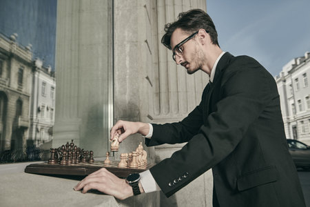 A serious man in a black business suit plays chess with his reflection, standing at the window of a building on the street of a big city. Business and political concept. Strategy and tactics.の写真素材