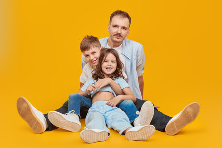 A happy father sits on the floor with his beloved son and daughter, arms outstretched cheerfully. Happy family concept. Full length portrait on a yellow studio background.の写真素材