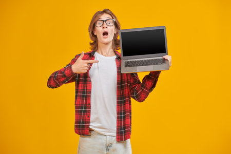 Emotional boy teenager schoolboy holds an open laptop in his hand and opens his mouth in surprise. Educational concept. Yellow studio background with copy space.の写真素材