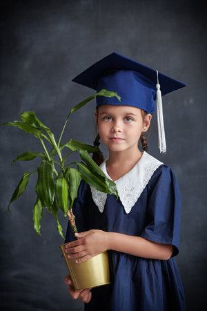 Education, biology. A little schoolgirl in a school uniform and an academic hat stands against the black chalkboard with a plant in her hands.の写真素材