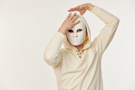 People and life roles. Portrait of a man in a white hoodie who hides his face behind a mask. Studio shot on a white background. Place for text.の写真素材