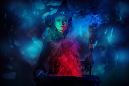 Halloween time. A beautiful young witch in a black dress and hat conjures over a cauldron on a dark...