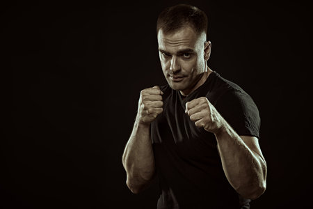 A strong, confident man with an athletic build stands in combat readiness, holding clenched fists in front of him. Studio portrait. Power sports, health and active life position. Fighter. Place for text.の写真素材
